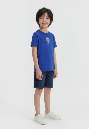 Garçon debout et souriant, portant un T-shirt bleu du Paris Saint-Germain, un short en jean foncé et des baskets blanches, sur un fond uni.