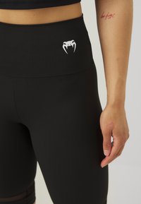 Sorte leggings med høy midje, med en hvit kobra logo på fronten og gjennomsiktige paneler langs sidene. Glatt tekstur.