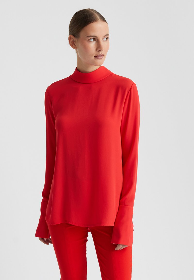 Rotes Langarmoberteil mit hohem Kragen, glatter Textur und legerem Schnitt. Verfügt über breite Bündchen und eine fließende Silhouette. Kombiniert mit roten Hosen.