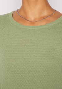 Maglione verde a maglia con scollo rotondo, tessuto testurizzato con un motivo sottile e finitura liscia e leggera. Accentuato da una catena d'argento.