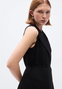 Μαύρο φόρεμα χωρίς μανίκια με βαθύ V λαιμόκοψη και πλισέ σχεδιασμό. Διαθέτει εφαρμοστή μέση και λεία υφή. Μινιμαλιστικό στυλ, χωρίς σχέδια.