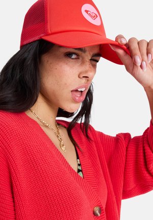 Rode meshpet met vooraan een logo, rond ontwerp en gebogen klep. Het model draagt een bijpassende rode cardigan met textuur en knoopdetails.