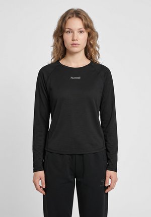 Ung kvinde med bølget hår iført en sort hummel langærmet trøje og sorte bukser, stående mod en ensfarvet lys baggrund.