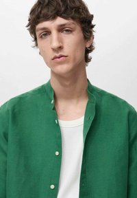 Green linen shirt with a stand-up collar, featuring white buttons and worn over a cream-colored t-shirt. Smooth texture and casual design.