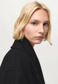 Zwarte wollen jas met een reverskraag, gemaakt van een gestructureerde stof. Model heeft kort blond haar en een neutrale gezichtsuitdrukking, kijkt naar links.