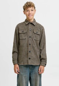 Camicia check con motivi marroni e beige, dotata di due tasche frontali, chiusura con bottoni e colletto, abbinata a pantaloni in denim blu.