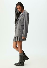 Blazer grigio in lana dal taglio sartoriale, indossato su una gonna grigia plissettata. Accompagnato da stivaletti neri alla caviglia e calze grigie al ginocchio.