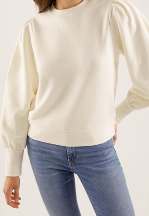 Kvinde iført en cremefarvet, langærmet sweater med puffede skuldre og tætsiddende manchetter, kombineret med blå jeans i denim.