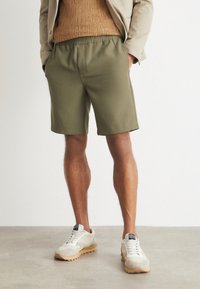 Olive green shorts made of smooth fabric, featuring an elastic waistband and side pockets, paired with light-colored sneakers.