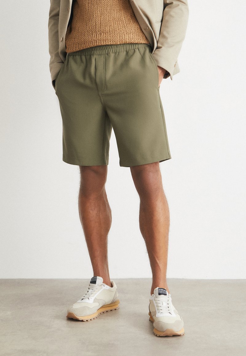 Olive green shorts made of smooth fabric, featuring an elastic waistband and side pockets, paired with light-colored sneakers.