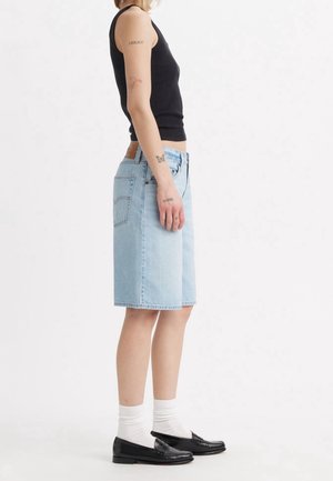 Persoon draagt een zwart tanktopje, lichtblauwe knielange spijkershorts, witte sokken en zwarte loafers, staand in profiel tegen een witte achtergrond.