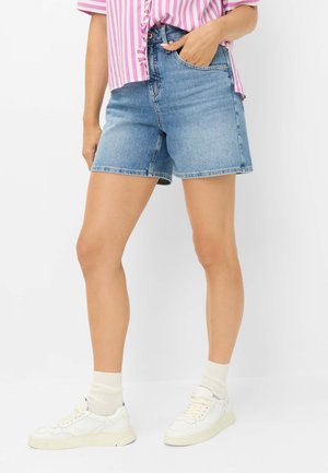 Person iført blå denimshorts i mellemlængde, hvide sneakers med hvide sokker og en pink og hvidstribet skjorte med hånden i lommen.