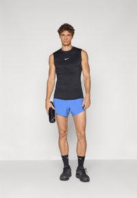 Schwarzes ärmelloses Sporttop mit weißem Logo, blaue Shorts, hält eine schwarze Wasserflasche, trägt schwarze Laufschuhe und Socken.
