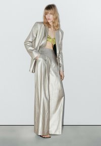 Metallic zilveren tailleurblazer gecombineerd met hoogcelde, wijde broek. Olijfgroen crop top zichtbaar onder de jas. Zachte textuur.