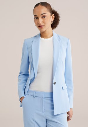 Vrouw met krullend haar in een lage knot, draagt gouden creolen, lichtblauw colbert en broek, wit geribd topje, poseert met één hand in de zak.