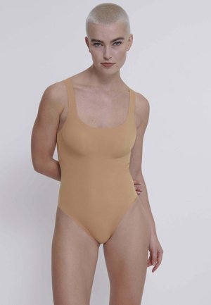 Persona con capelli biondi corti che indossa un costume intero beige senza maniche, in piedi con una mano dietro la schiena su uno sfondo uniforme.
