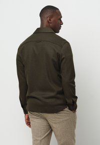 Camicia tessuta verde scuro con colletto, maniche lunghe e polsini con bottone; presenta un orlo dritto sul retro e una texture sottile.