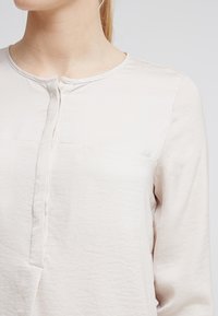 Camicia a maniche lunghe di un leggero beige, caratterizzata da un collo rotondo, una parziale apertura con bottoni e una sottile trama sgualcita. Dettaglio della tasca frontale assessabile.