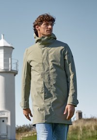 Parka imperméable vert clair avec un col montant et une capuche, dotée de manches longues et d'une coupe droite, portée par-dessus un jean bleu à l'extérieur.