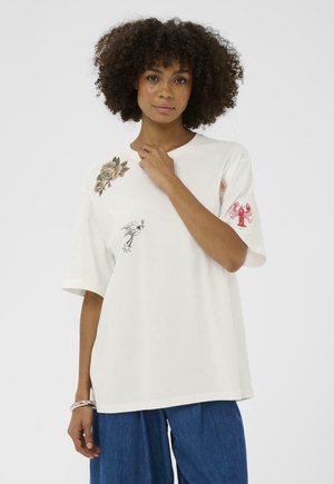 Jeune femme aux cheveux bouclés portant un t-shirt blanc oversize orné de motifs floraux, d'oiseaux et de homards, ainsi qu'un pantalon large bleu.
