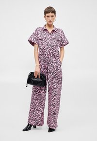 Rózsaszín leopárd mintás overál rövid ujjakkal, széles lábakkal és megkötött derekú kivitelben. Egy kis fekete kézitáskával és hegyes orrú fekete csizmával párosítva.