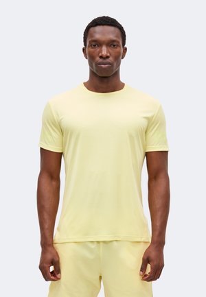 Uomo con pelle scura che indossa una camicia a maniche corte giallo chiaro e pantaloncini abbinati, in piedi davanti a uno sfondo chiaro e uniforme.