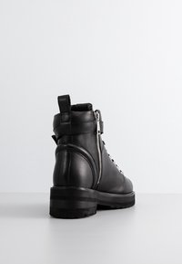 Bottine en cuir noir avec fermeture éclair latérale argentée, lacets à l'avant, languette de tirage et semelle en caoutchouc épaisse, vue de dos et de côté.