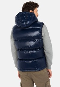 Navy blauer Puffer-Weste mit glänzender Oberfläche, Kapuze und gestepptem Design. Über einem hellgrauen Langarmshirt getragen, mit sichtbaren Nähten und Detail an den Nähten.