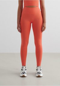 Leggings a vita alta color corallo in tessuto elasticizzato con una superficie liscia. Abbinati a sneaker bianche e nere con dettagli a contrasto.