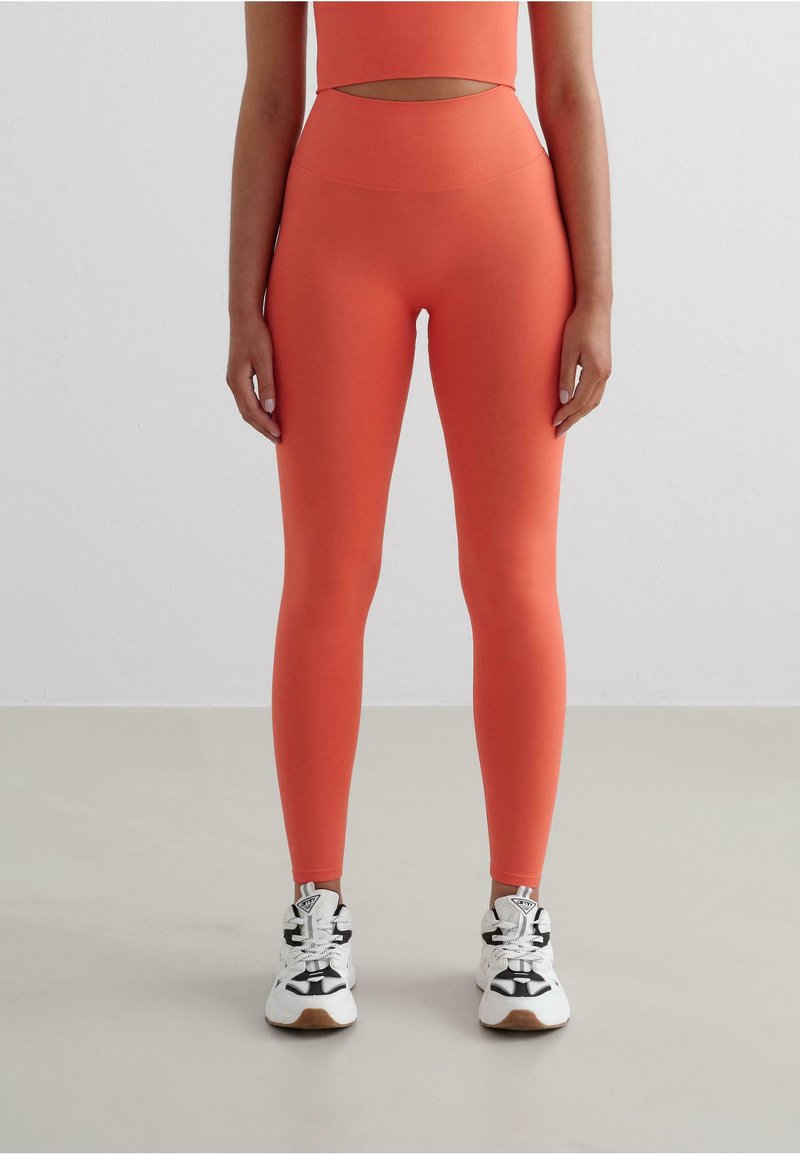 Leggings a vita alta color corallo in tessuto elasticizzato con una superficie liscia. Abbinati a sneaker bianche e nere con dettagli a contrasto.