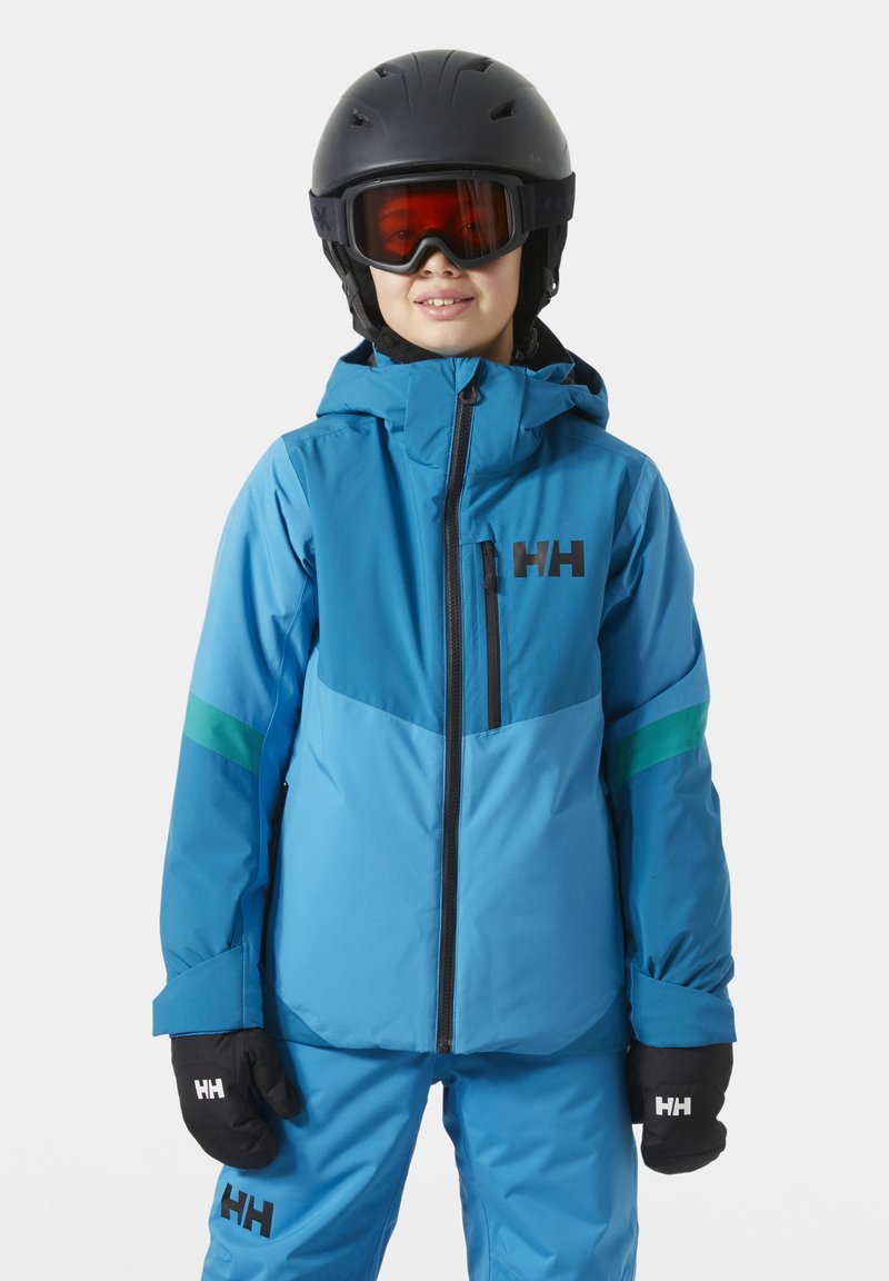 Barn iført blå skijakke og bukser, sort hjelm, skibriller med rødt skær og sorte handsker med mærkelogo på brystet og handskerne.