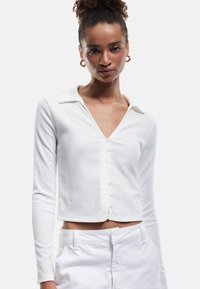 Witte lange mouwen crop top van een textuurstof, met een V-halslijn en knoopsluiting aan de voorkant. Met kraag ontwerp.