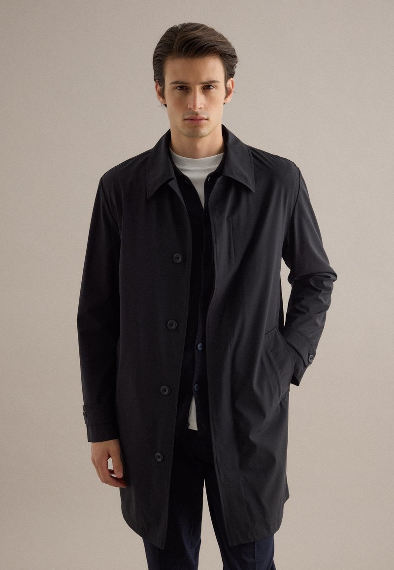 Giovane uomo in piedi, indossa un cappotto nero con bottoni sopra una camicia bianca, con una mano nella tasca del cappotto, sfondo neutro.