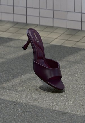 En enkelt mørkelilla åben tå højhælet sandal stående oprejst på en grå belægning nær en flisevæg.