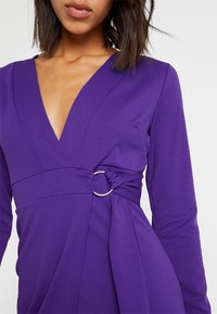 Robe violette à col en V en tissu lisse, avec un détail torsadé agrémenté d'un anneau argenté à la taille et des manches longues.