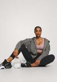 Graue Zip-Hoodie mit pinken Akzenten, getragen über einem hellpinken Sport-BH und schwarzen Leggings. Schwarze Sneakers. Eine klare Wasserflasche in der Nähe.