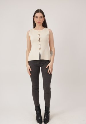 Jonge vrouw met lang donker haar die een mouwloze crèmekleurige top met knopen draagt, donkere strakke jeans, zwarte enkellaarzen en een bloemen-tatoeage op haar linker onderarm.