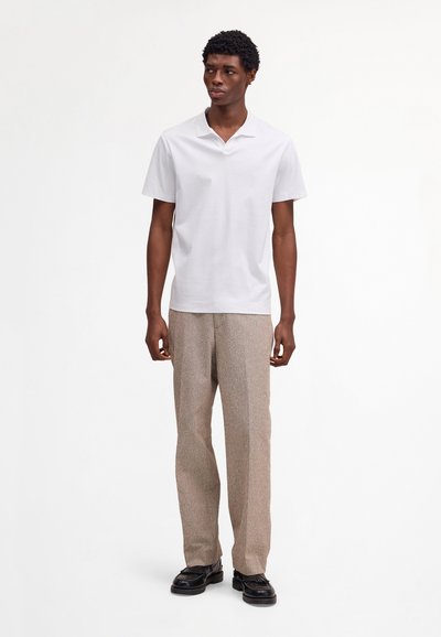 Man standing wearing a plain white short-sleeve polo shirt, beige textured pants, and black leather shoes on a white background.