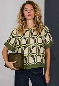 Lõigatud top kreemjas ja rohelises geomeetrilises mustris mustade aktsentidega. Modell hoiab rohelisest nahast kotikest, mis sobib tumedate teksadega.