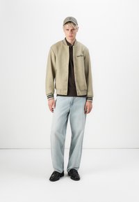 Beige wool varsity jacket with black and white striped cuffs, paired with a dark brown polo and light blue relaxed-fit jeans. Black shoes.