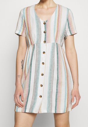 Donna che indossa un vestito a maniche corte con bottoni, caratterizzato da strisce verticali pastello in turchese, rosa, giallo e marrone su uno sfondo bianco.