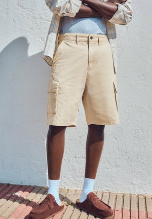 Uomo con pantaloncini cargo beige, calzini azzurri, scarpe marroni slip-on e camicia a quadri, con le braccia incrociate contro un muro bianco.