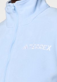 Hele sinine fliisist lukuga jakk, millel on kõrge krae ja tikitud "TERREX" logo. Sile tekstuur ja lukuga kinnituse detail.