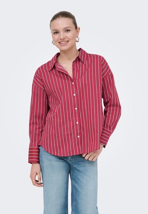 Lächelnde Frau trägt ein rotes langärmeliges Hemd mit weißen vertikalen Streifen und blaue Jeans, steht mit einer Hand in der Tasche.