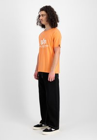 Oranges T-Shirt mit kurzen Ärmeln und weißem Grafiktext, schwarze weite Hose und schwarze Sneakers mit weißen Sohlen. Lockige Haare, die das Gesicht umrahmen.