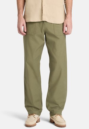 Mann trägt olivgrüne Hose mit geradem Bein, beiges Hemd und hellbraune Schnürschuhe vor einem schlichten weißen Hintergrund.