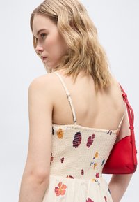 Νεαρή γυναίκα με κυματιστά ξανθά μαλλιά, ντυμένη με ένα κρεμ φόρεμα με floral σχέδιο και λεπτές τιράντες, κρατώντας μια κόκκινη τσάντα ώμου.