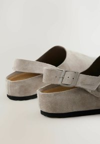 Mules en suede gris avec une large sangle et une boucle rectangulaire, présentant une semelle intérieure en liège et une semelle en caoutchouc noir texturé.