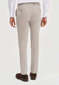 Jasnobeżowe spodnie o subtelnej fakturze, z tylnymi kieszeniami zapinanymi na guziki. Noszone z brązowymi zamszowymi butami. Dopasowany, slim-fit fason.