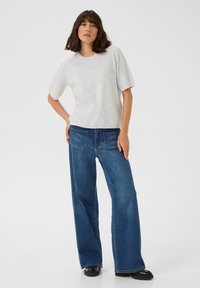 Un pull texturé gris à manches courtes associé à un jean bleu à larges jambes, doté de poches arrière et de chaussures noires épaisses.